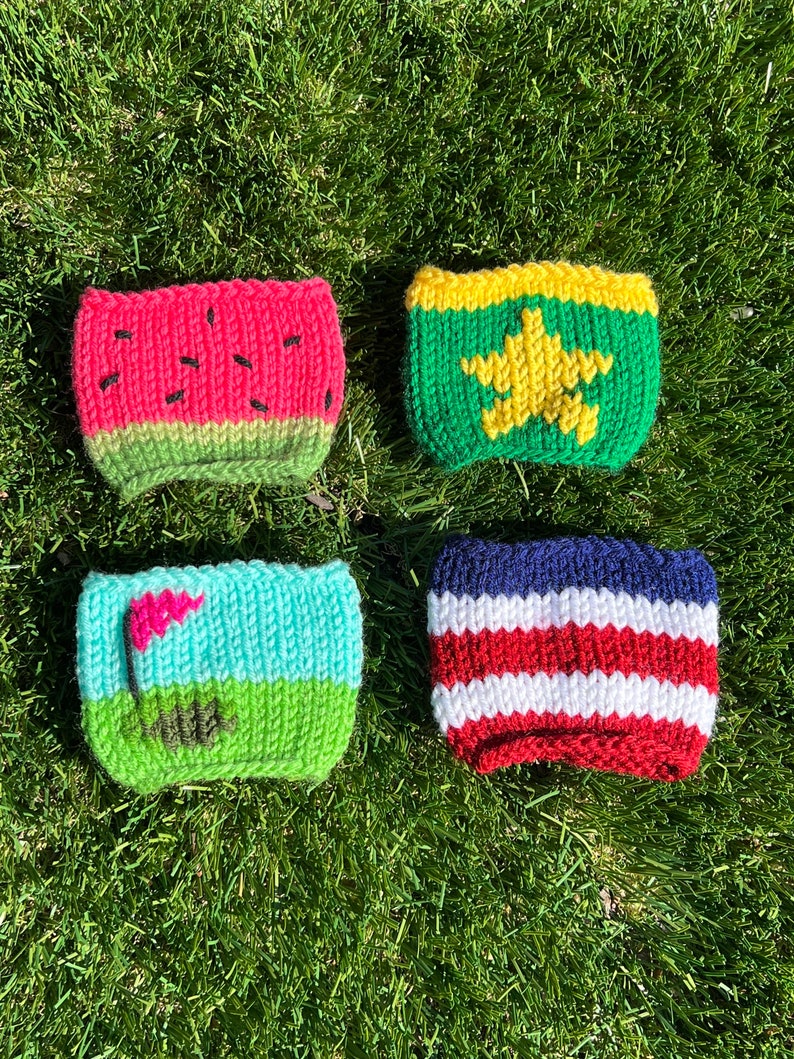 May include: Four knitted cup cozies with different designs. The first is watermelon, the second is a green and yellow star, the third is a golf course, and the fourth is red, white, and blue stripes.