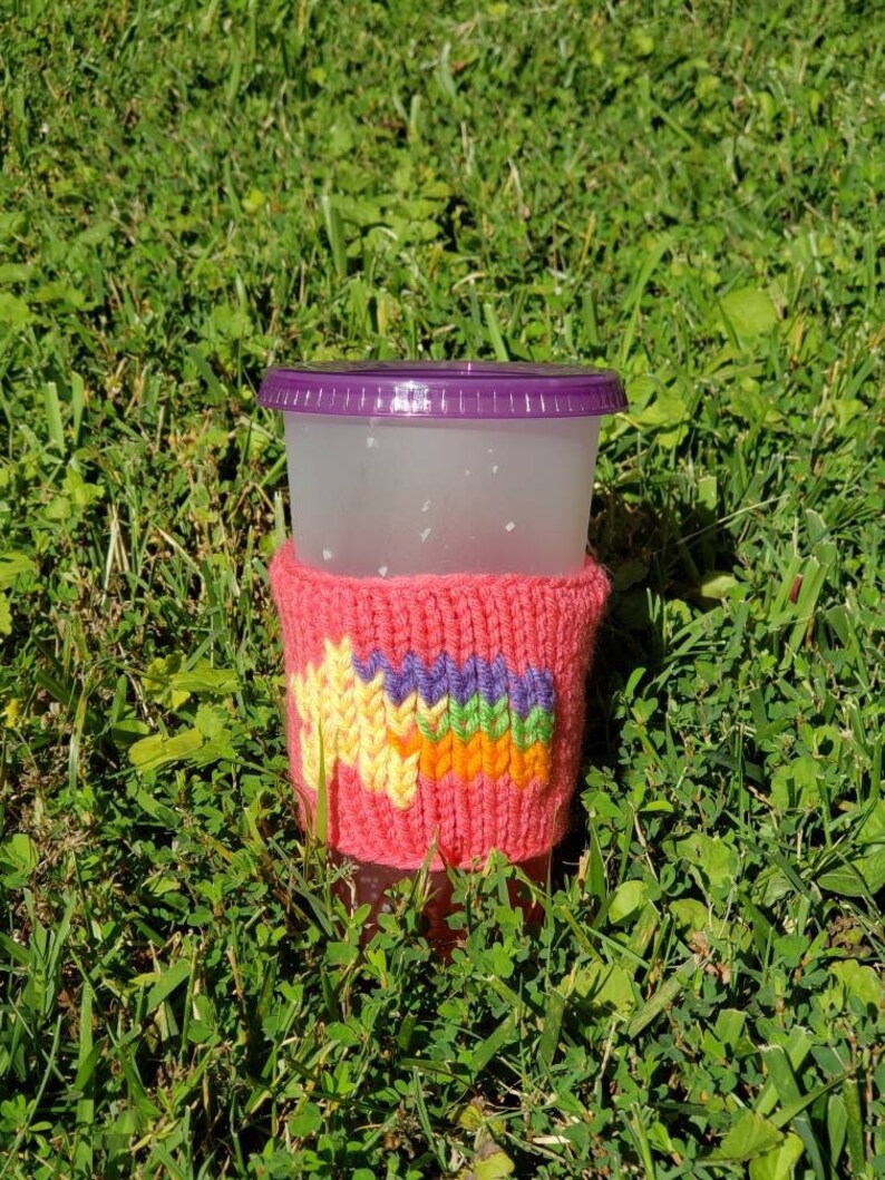 May include: A pink knitted cup cozy with a rainbow design. The cozy is on a clear plastic cup with a purple lid. The cup is sitting on green grass.