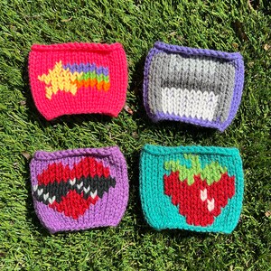 May include: Four knitted cup cozies in different colors and designs. The first is pink with a yellow star and rainbow. The second is purple with a gray and white stripe. The third is purple with a red heart. The fourth is teal with a red strawberry.