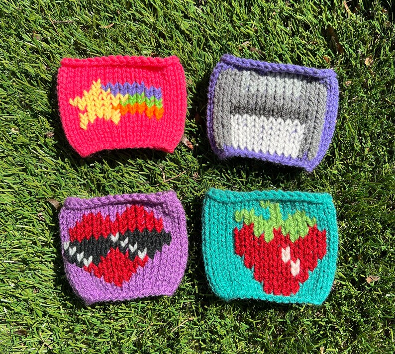 May include: Four knitted cup cozies in different colors and designs. The first is pink with a yellow star and rainbow. The second is purple with a gray and white stripe. The third is purple with a red heart. The fourth is teal with a red strawberry.