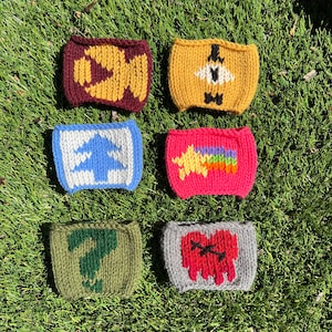 May include: Six knitted cup cozies with different designs. The cozies are in various colors, including red, yellow, blue, green, and gray. Each cozy features a unique design, such as a question mark, a heart, a rainbow, and a pine tree.