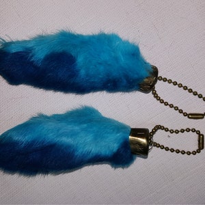 May include: Two blue rabbit foot keychains with gold metal rings and clasps. The rabbit feet are dyed a gradient of blue.