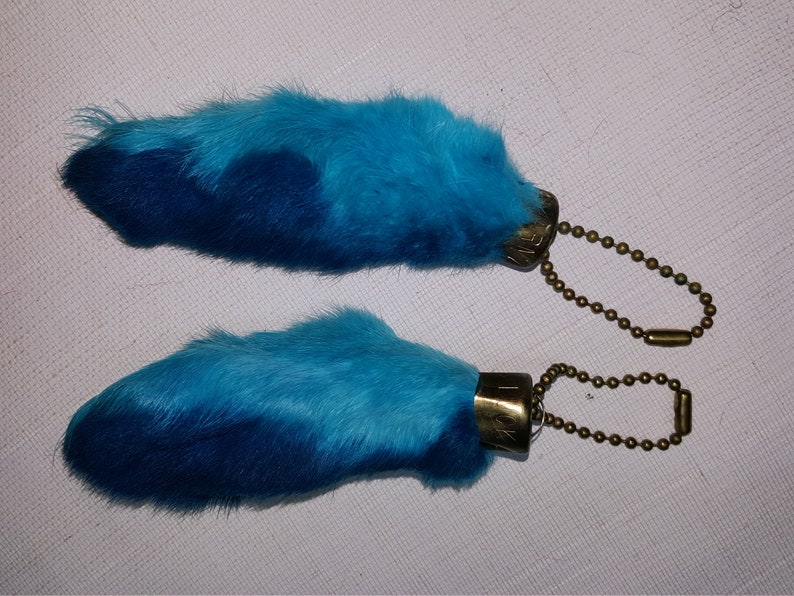 May include: Two blue rabbit foot keychains with gold metal rings and clasps. The rabbit feet are dyed a gradient of blue.