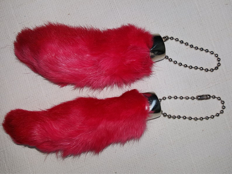 May include: Two pink rabbit foot keychains with silver metal rings and chains. The rabbit feet are made of fur.