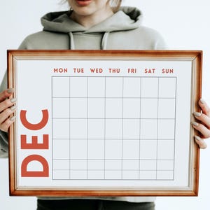 Könnte beinhalten: Ein gerahmter Kalender für Dezember, gehalten von einer Person. Das Kalenderraster ist leer, mit den Wochentagen oben in Rot. Der Monat "DEC" ist vertikal in Rot auf der linken Seite gedruckt. Der Rahmen hat eine warme braune Farbe.