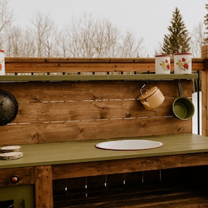 May include: A wooden play kitchen with a green countertop and a brown wooden base. The kitchen has a stovetop, a sink, and a shelf with various kitchen utensils. The kitchen is set up outdoors on a wooden deck.