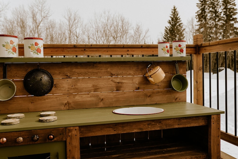May include: A wooden play kitchen with a green countertop and a brown wooden base. The kitchen has a stovetop, a sink, and a shelf with various kitchen utensils. The kitchen is set up outdoors on a wooden deck.