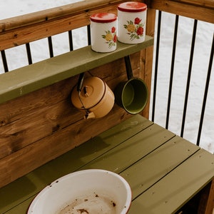 May include: A wooden play kitchen with a green countertop and a white enamel sink. There are two red and white floral canisters on a shelf above the sink, and a yellow kettle and a green pot hanging from hooks.