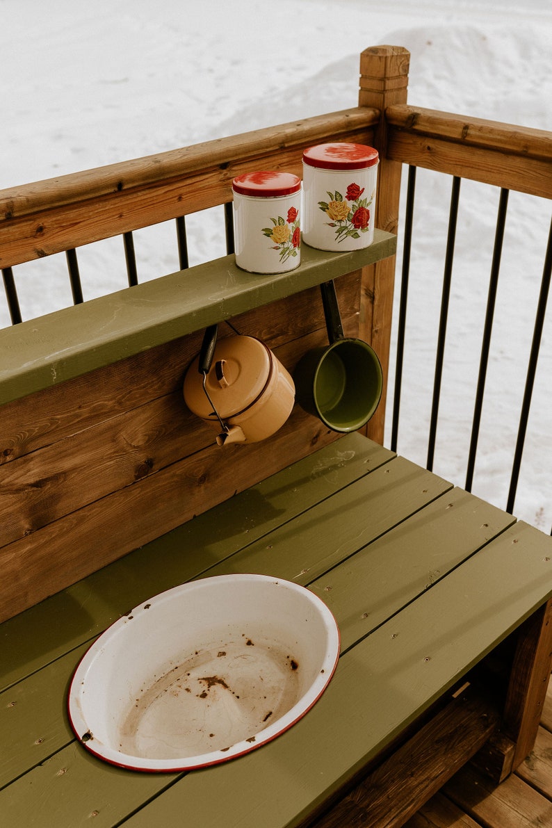 May include: A wooden play kitchen with a green countertop and a white enamel sink. There are two red and white floral canisters on a shelf above the sink, and a yellow kettle and a green pot hanging from hooks.