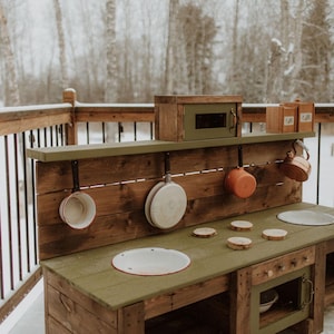Puede incluir: Una cocina de juguete de madera con una encimera verde, un horno peque&ntilde;o y un estante con ganchos para colgar ollas y sartenes. La cocina est&aacute; hecha de madera natural y tiene un aspecto r&uacute;stico.