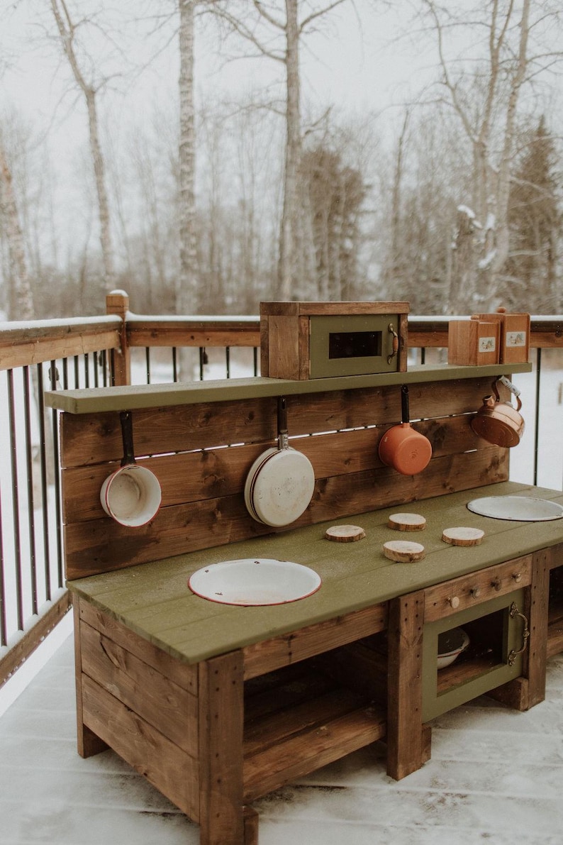 Puede incluir: Una cocina de juguete de madera con una encimera verde, un horno peque&ntilde;o y un estante con ganchos para colgar ollas y sartenes. La cocina est&aacute; hecha de madera natural y tiene un aspecto r&uacute;stico.