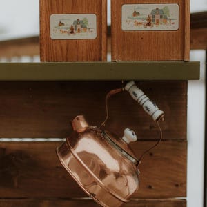 Puede incluir: Dos cajas de madera con un dise&ntilde;o de escena campestre se sientan en un estante de madera. Una tetera de cobre con un asa blanca cuelga del estante.