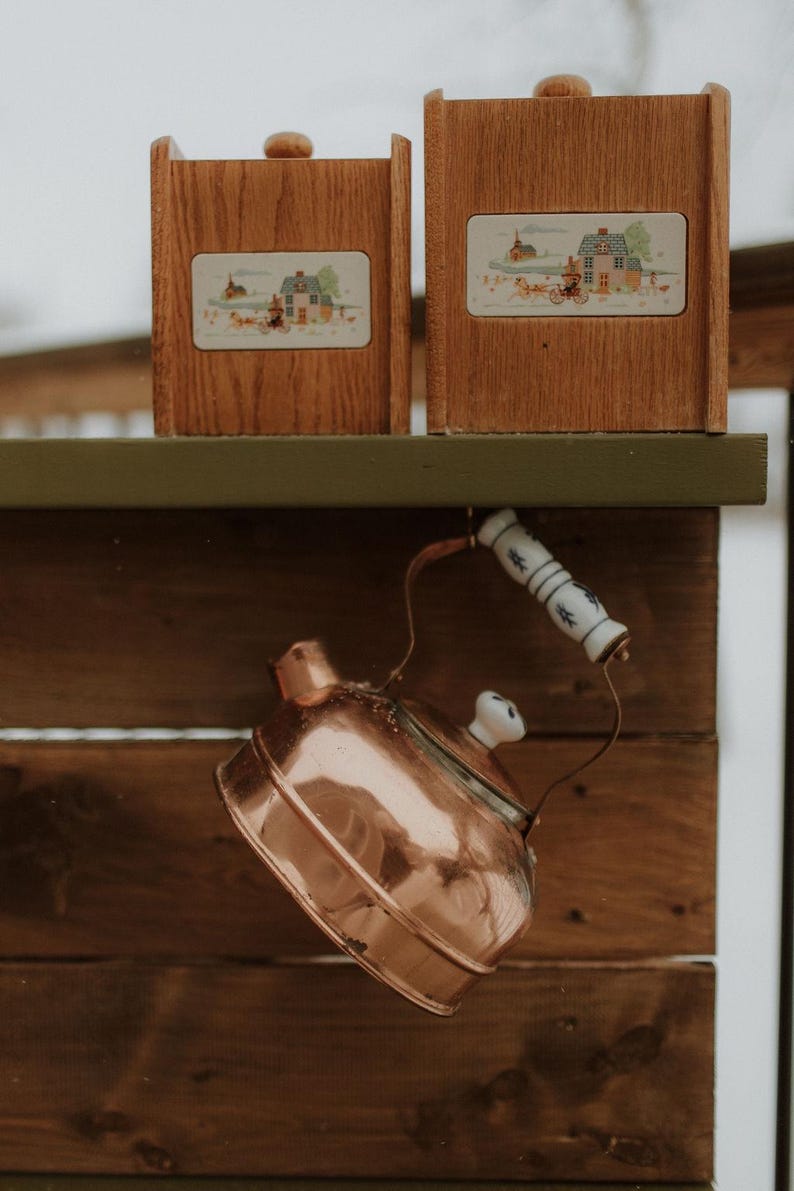 Puede incluir: Dos cajas de madera con un dise&ntilde;o de escena campestre se sientan en un estante de madera. Una tetera de cobre con un asa blanca cuelga del estante.