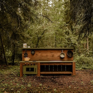 May include: A wooden play kitchen set with a green oven, a brown countertop, and a shelf. The kitchen is set up in a forest setting.