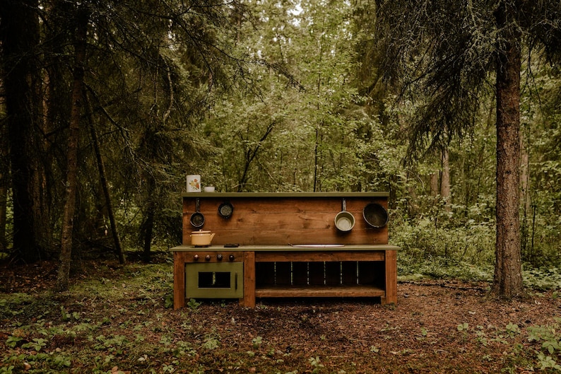 May include: A wooden play kitchen set with a green oven, a brown countertop, and a shelf. The kitchen is set up in a forest setting.