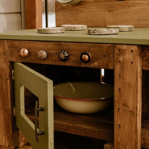 May include: A wooden play kitchen with a green painted countertop and a green cabinet door. The countertop has four round wooden stovetop burner covers and four decorative knobs. The cabinet has a black handle and a white bowl inside.