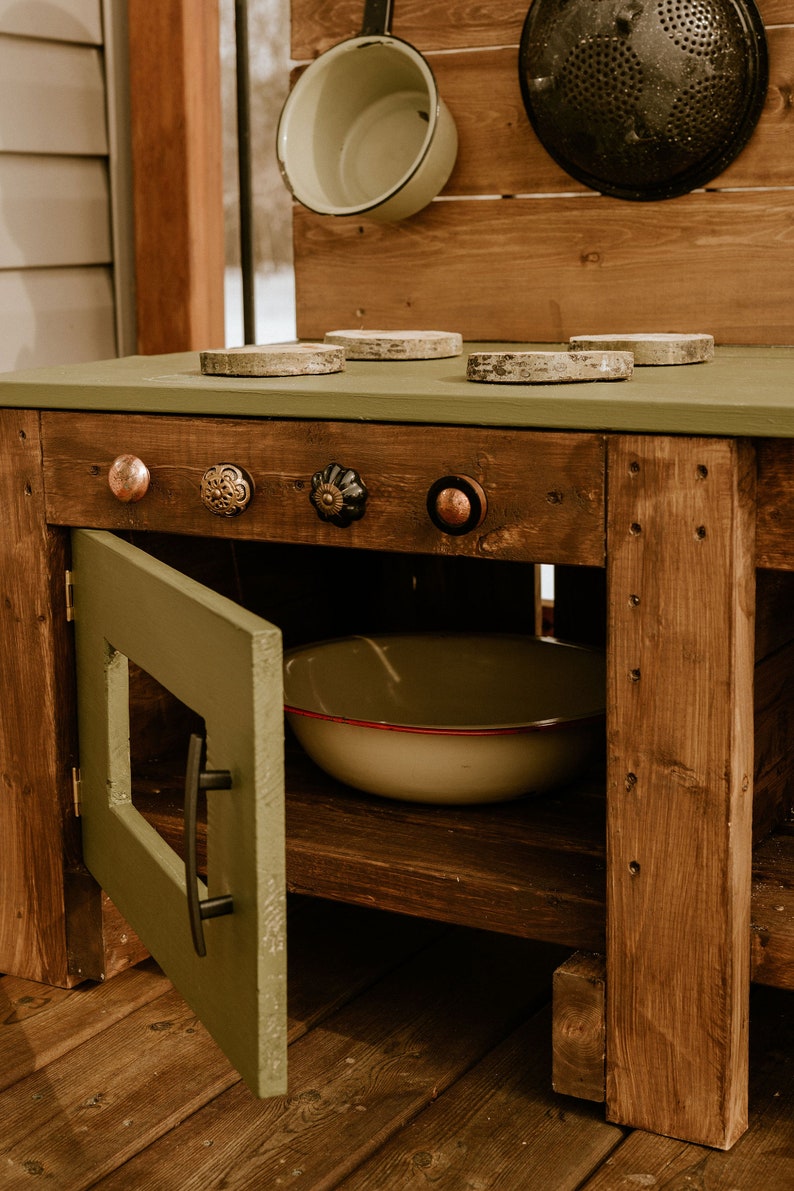 May include: A wooden play kitchen with a green painted countertop and a green cabinet door. The countertop has four round wooden stovetop burner covers and four decorative knobs. The cabinet has a black handle and a white bowl inside.