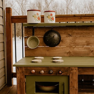 May include: A wooden play kitchen with a green countertop and a green oven door. The kitchen has a wooden shelf with two white canisters with floral designs and a black colander hanging below. There are four wooden circles on the countertop, and five knobs on the oven door.