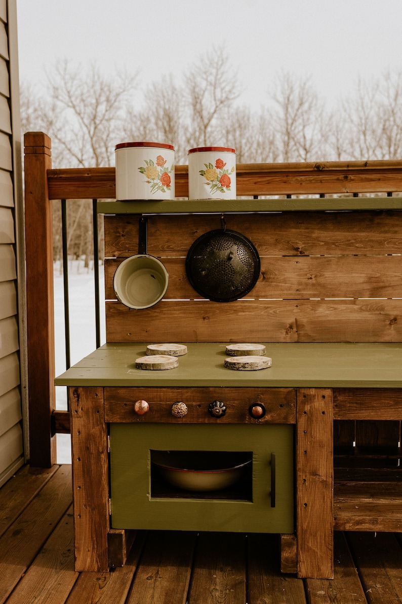 May include: A wooden play kitchen with a green countertop and a green oven door. The kitchen has a wooden shelf with two white canisters with floral designs and a black colander hanging below. There are four wooden circles on the countertop, and five knobs on the oven door.