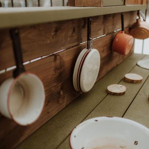 Puede incluir: Una cocina de juguete de madera con una variedad de ollas y sartenes de esmalte blanco y rojo colgadas de ganchos. La encimera de la cocina es de madera y tiene una superficie verde claro. Hay varios c&iacute;rculos de madera en la encimera.