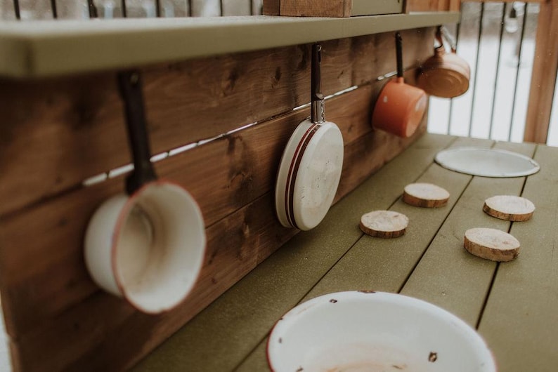 Puede incluir: Una cocina de juguete de madera con una variedad de ollas y sartenes de esmalte blanco y rojo colgadas de ganchos. La encimera de la cocina es de madera y tiene una superficie verde claro. Hay varios c&iacute;rculos de madera en la encimera.
