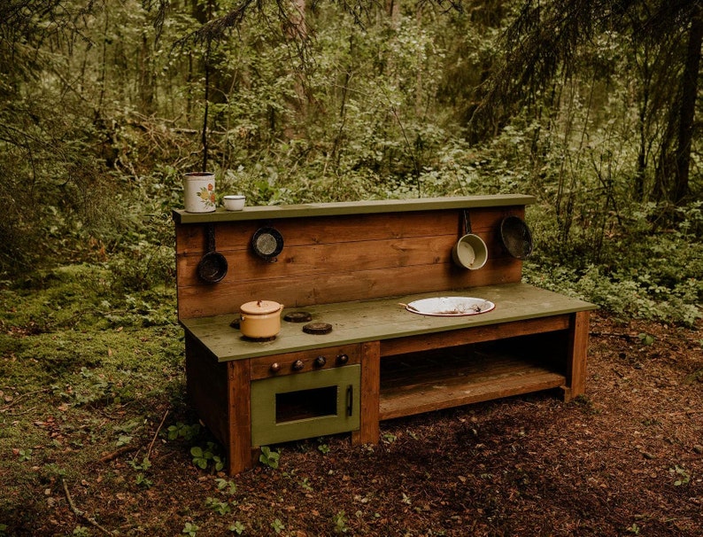 May include: A wooden play kitchen set with a green countertop, a small oven, and a sink. The kitchen is set up in a forest setting with green moss and trees in the background.