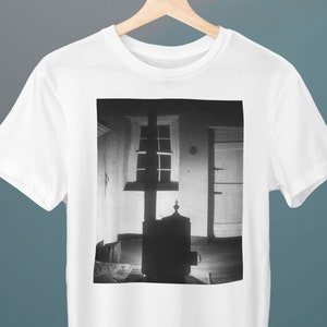 Puede incluir: Fotografía en blanco y negro de una habitación con una ventana, una puerta y una estufa de leña. La estufa está en el centro de la imagen y está silueteada contra la luz de la ventana.