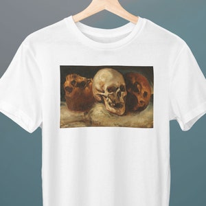 May include: A white t-shirt with a print of a painting of three human skulls. The skulls are arranged in a row, with the largest skull in the center. The skulls are painted in a realistic style, with details such as the teeth and eye sockets.