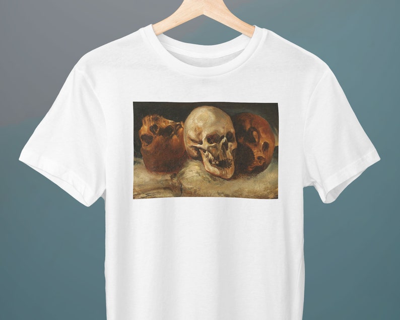 May include: A white t-shirt with a print of a painting of three human skulls. The skulls are arranged in a row, with the largest skull in the center. The skulls are painted in a realistic style, with details such as the teeth and eye sockets.