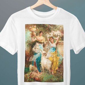 以下が含まれることがあります： 白いTシャツに、庭の風景の中にいる3人の女性を描いたヴィンテージの絵がプリントされています。女性たちは、ゆったりとしたドレスを着て、花を持っています。白いヤギが画像の中央に立っています。
