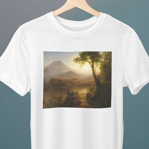 May include: White t-shirt with a print of a landscape painting. The painting depicts a mountain range, a lake, and a large tree in the foreground. The sky is a light blue with clouds.