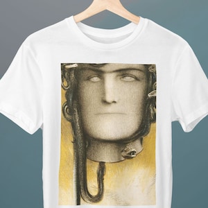 Op de afbeelding: Wit T-shirt met een print van een schilderij van een persoon met slangen om zijn hoofd. De persoon heeft een bleek gezicht en witte ogen. De achtergrond is geel en bruin.