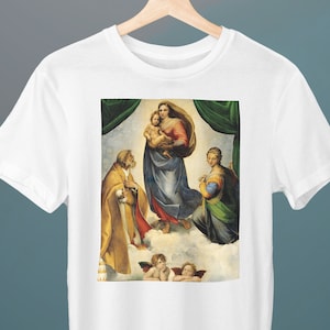 Puede incluir: Una camiseta blanca con una impresión de una pintura renacentista que representa a la Virgen y el Niño con San José y dos ángeles. La pintura está sobre un fondo de cielo azul y nubes.