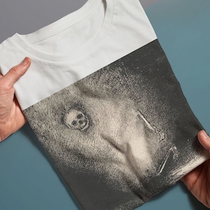 Puede incluir: Una camiseta blanca con un estampado gr&aacute;fico en blanco y negro de una calavera y una figura sosteniendo un cuchillo.