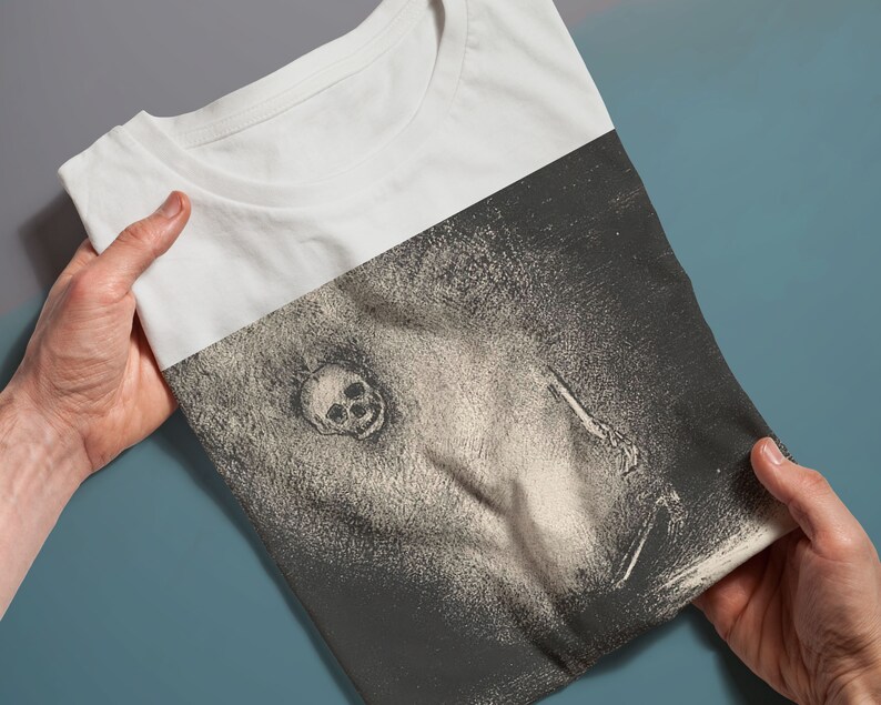 Puede incluir: Una camiseta blanca con un estampado gr&aacute;fico en blanco y negro de una calavera y una figura sosteniendo un cuchillo.