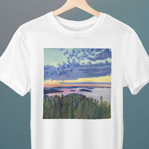 May include: White t-shirt with a print of a painting of a lake and sky with clouds. The sky is a light blue with white clouds and a yellow sunset. The lake is a light blue with green trees on the shore.