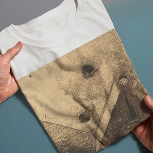 May include: A white t-shirt with a vintage-style drawing of a person in a hat and a cloak. The drawing is in shades of brown and beige.