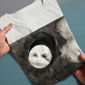 May include: White t-shirt with a black and white illustration of a child's face in a circle, surrounded by clouds.