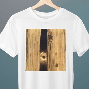 Puede incluir: Camiseta blanca con un diseño cuadrado de vetas de madera. El diseño presenta tablas de madera verticales en tonos marrón y negro, con nudos y detalles de vetas visibles. La camiseta se muestra en una percha de madera.