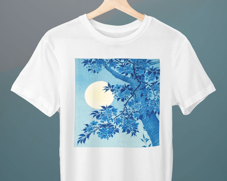 K&ouml;nnte beinhalten: Wei&szlig;es T-Shirt mit einem blauen und wei&szlig;en Druck eines Astes mit blauen Bl&uuml;ten und einem Vollmond im Hintergrund.