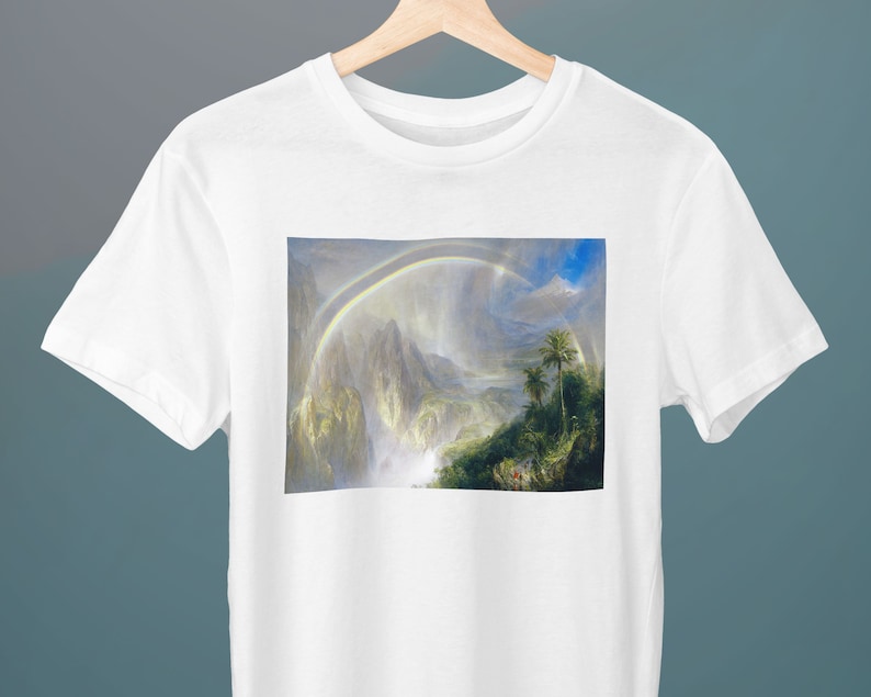 May include: A white t-shirt featuring a print of a painting depicting a lush green landscape with a waterfall and a rainbow in the sky.