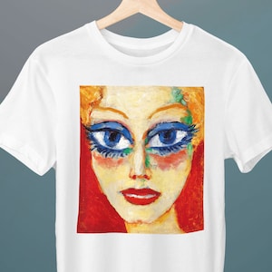 Puede incluir: Camiseta blanca con una pintura colorida de la cara de una mujer con ojos azules y labios rojos. La pintura está en un marco cuadrado en la parte delantera de la camiseta.