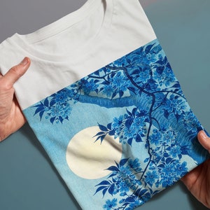 K&ouml;nnte beinhalten: Wei&szlig;es T-Shirt mit einem blauen und wei&szlig;en Blumendruck mit einem gro&szlig;en Mond. Der Druck erinnert an japanische Kunst.
