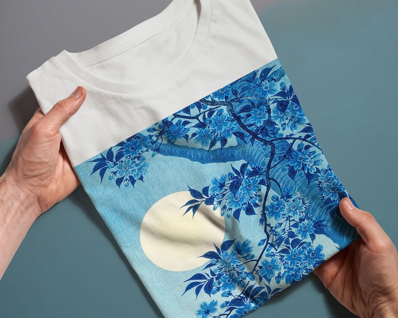 K&ouml;nnte beinhalten: Wei&szlig;es T-Shirt mit einem blauen und wei&szlig;en Blumendruck mit einem gro&szlig;en Mond. Der Druck erinnert an japanische Kunst.