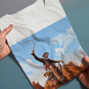 May include: A white t-shirt with a colorful image of a cowboy riding a horse and holding a whip against a blue sky with white clouds.