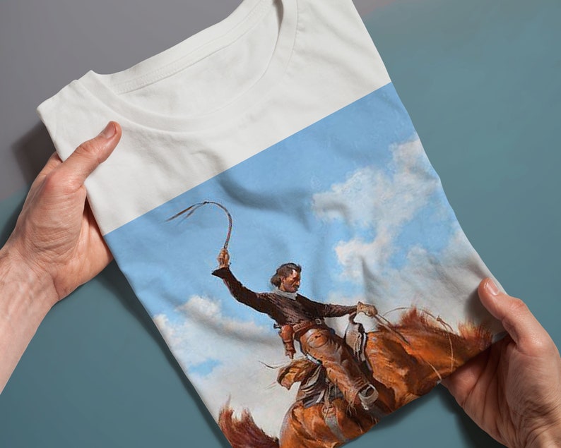 May include: A white t-shirt with a colorful image of a cowboy riding a horse and holding a whip against a blue sky with white clouds.
