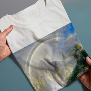 May include: A white t-shirt featuring a colourful rainbow and mountain landscape print. The print depicts a double rainbow over a verdant valley with mountains in the background.