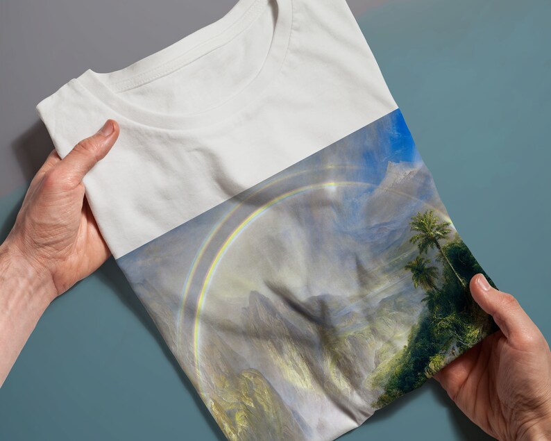 May include: A white t-shirt featuring a colourful rainbow and mountain landscape print. The print depicts a double rainbow over a verdant valley with mountains in the background.