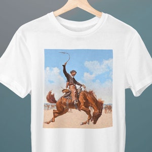 May include: A white t-shirt with a vintage illustration of a cowboy riding a bucking bronco. The cowboy is wearing a brown hat, a brown shirt, and brown pants. The horse is brown and white. The background is a blue sky with white clouds.