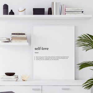 May include: A white framed print with the definition of self-love. The print is hanging on a white wall above a white shelf with books and decorative objects. A potted palm plant is in the corner of the image.